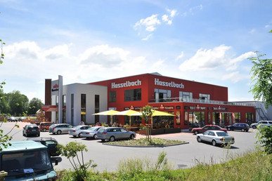 Das Bild zeigt ein modernes Gebäude mit roter Fassade, auf dem "Hasselbach" steht. Es gibt eine große Terrasse mit Sonnenschirmen und zahlreiche parkende Autos auf dem Parkplatz.