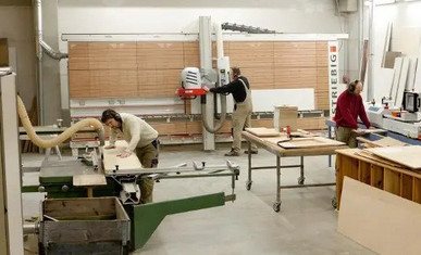 In einer Werkstatt arbeiten zwei Handwerker an Holzprojekten. Der erste Handwerker verwendet eine Maschine zum Schneiden von Holzplatten.
