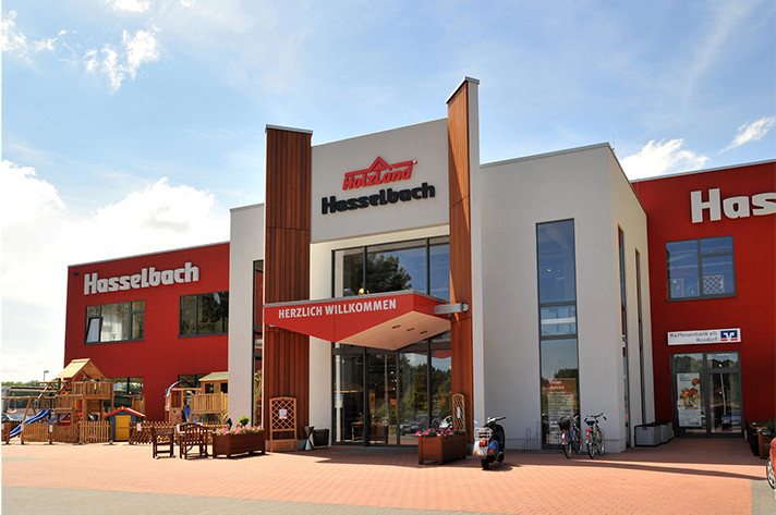 Das Bild zeigt den Haupteingang eines Geschäfts mit dem Namen "Hasselbach". Über dem Eingangsbereich steht "HERZLICH WILLKOMMEN". Das Gebäude hat große Fenster, eine moderne Architektur, umgeben von einem gepflasterten Bereich.