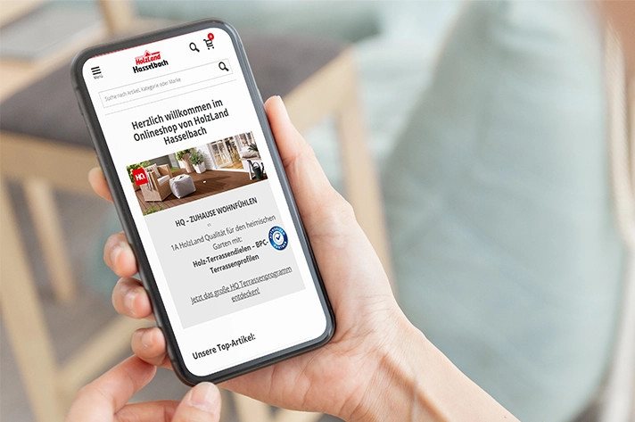Eine Person hält ein Smartphone in der Hand, auf dem der Online-Shop von HolzLand Hasselbach zu sehen ist.